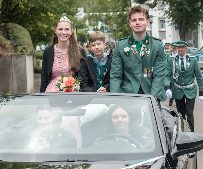 Eine junge Frau mit Tiara und Blumen, ein Junge und ein junger Mann in grüner Schützenuniform mit Orden stehen hinter einem schwarzen Cabriolet beim Festumzug.
