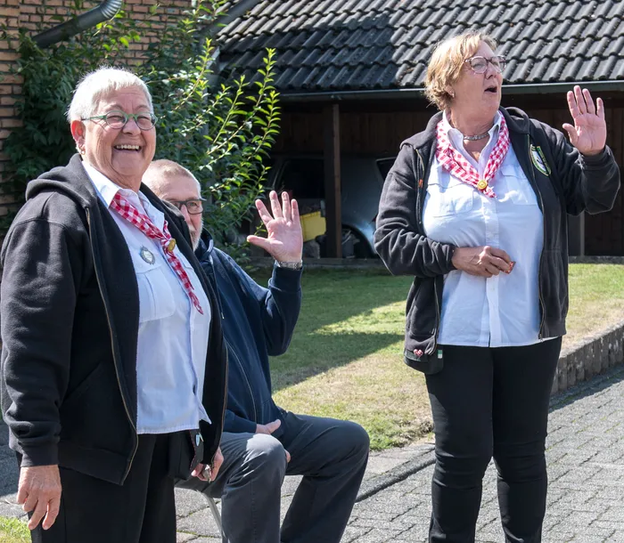 Zwei Frauen und ein Mann mit rot-weiß karierten Schals lachen und winken bei einem Festumzug.