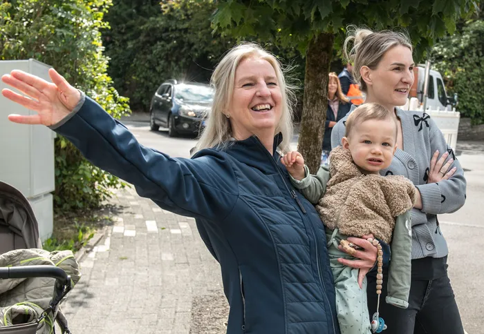 Eine lächelnde Frau in blauer Jacke winkt, während eine andere Frau ein Kleinkind hält.