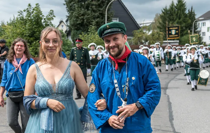 Ein Mann in blauer Jacke, grüner Kappe und rotem Halstuch sowie eine Frau in einem hellblauen Kleid vor einem Schützenfest-Umzug.
