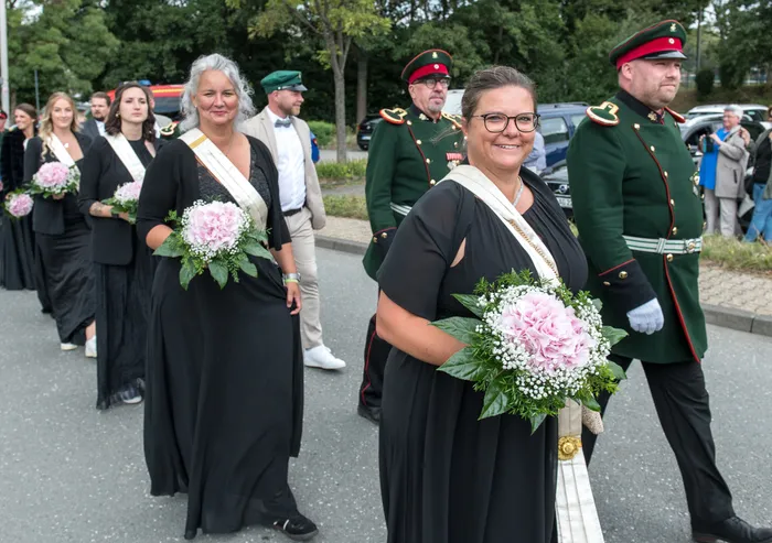 Frauen in schwarzen Kleidern mit weißen Schärpen und Blumensträußen ziehen neben Männern in grünen Schützenuniformen bei einem Festumzug durch Kierspe.