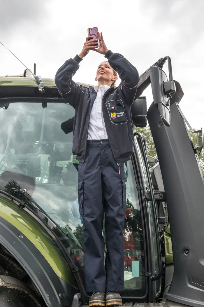 Eine Frau in einer dunkelblauen Feuerwehr-Uniform steht auf einem grünen Traktor und fotografiert mit einem rosa Smartphone nach oben.