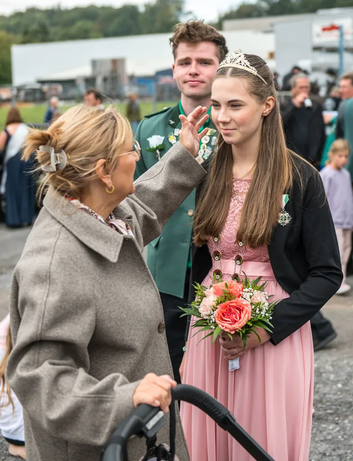 Eine Frau berührt das Gesicht einer jungen Frau in einem rosa Kleid mit Tiara und Blumenstrauß. Ein junger Mann in grüner Uniform steht dahinter.