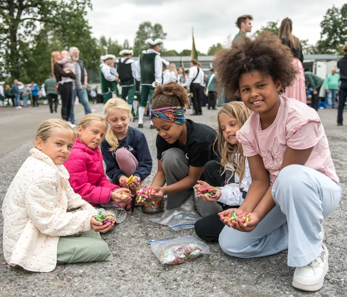 Sechs Mädchen sitzen auf dem Boden und halten bunte Süßigkeiten. Im Hintergrund sind Menschen in traditionellen Trachten bei einem Schützenfest zu sehen.