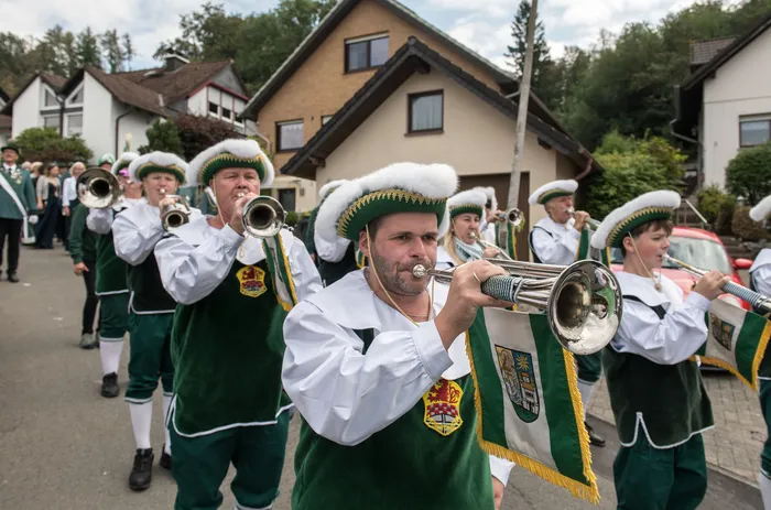 Mitglieder in grünen und weißen Uniformen mit Federhüten spielen Blasinstrumente bei einem Festumzug.