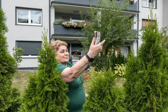Eine Frau in einem grünen T-Shirt hält ihr Smartphone hoch, um ein Foto vor einem Wohngebäude und grünen Büschen aufzunehmen.
