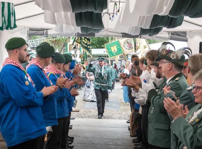 Ein Mann in grüner Festuniform geht durch einen Spalier aus applaudierenden Menschen in blauen und grünen Uniformen unter einem Festzelt.