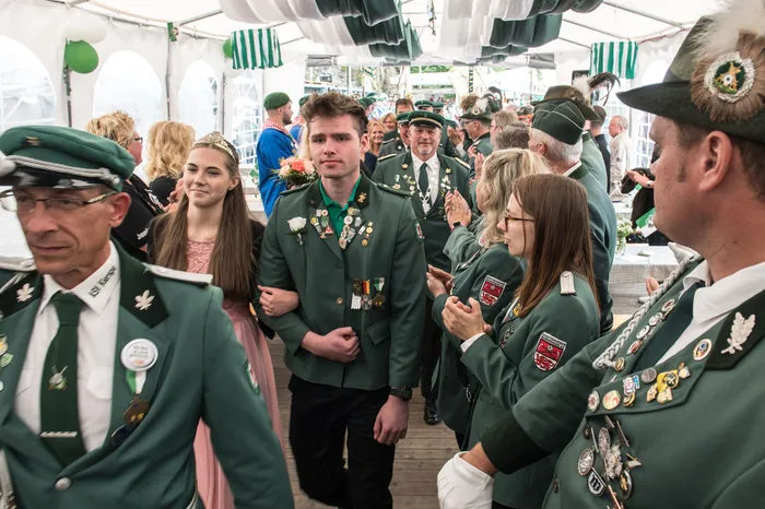 Personen in grünen Schützenuniformen mit Medaillen und eine junge Frau in einem rosa Kleid gehen durch ein Festzelt.