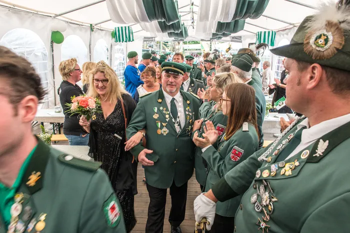 Ein Mann in grüner Uniform mit vielen Orden und eine Frau mit Blumenstrauß stehen inmitten von Menschen in grünen Trachten in einem Festzelt.