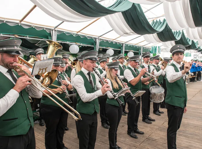 Musiker in grünen Westen und grauen Mützen spielen Blasinstrumente unter einem grün-weißen Festzelt.