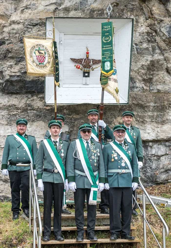 Männer in grünen Schützenuniformen mit weißen Schärpen und Orden stehen vor einer Felswand mit Vereinsfahnen.