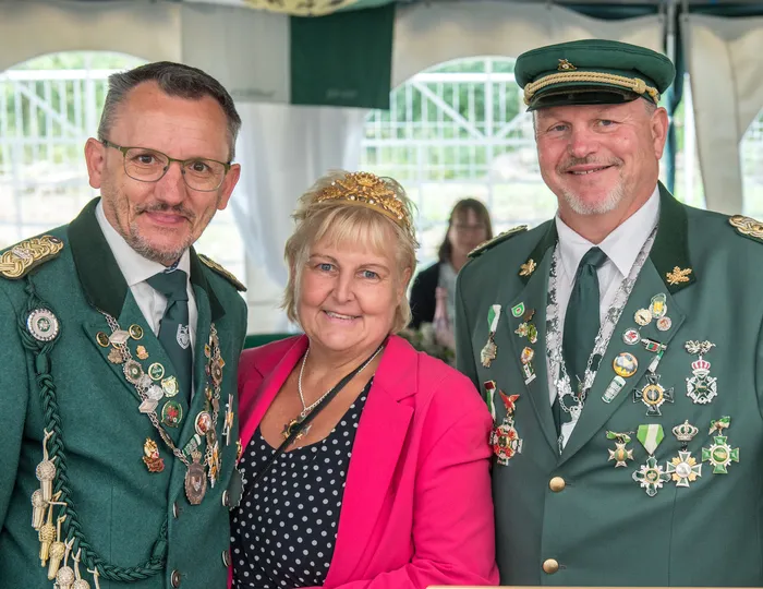 Zwei Männer in grünen Schützenuniformen mit zahlreichen Orden und eine Frau mit goldener Krone und pinkem Blazer.