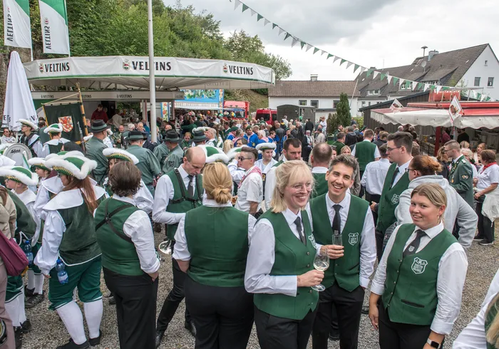 Menschen in traditioneller Kleidung mit grünen Westen und Hüten stehen bei einem Schützenfest vor Zelten und Ständen.
