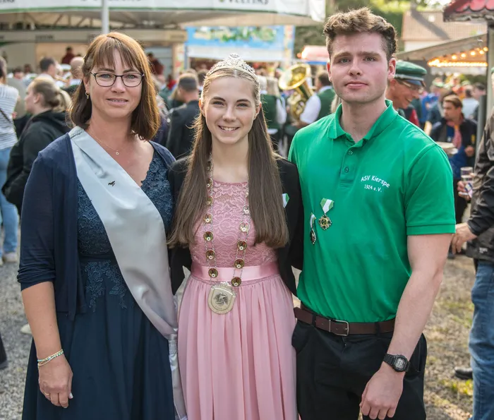 Eine junge Frau mit Tiara und Medaille in einem rosa Kleid steht zwischen einer Frau mit silberner Schärpe und einem Mann im grünen ASV Kierspe Poloshirt.