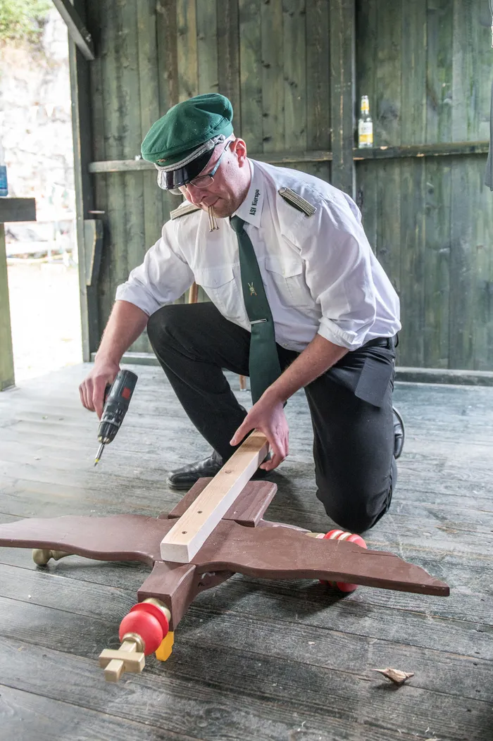 Ein Mann in weißer Uniform mit grüner Krawatte und Mütze kniet auf einem Holzboden und bearbeitet mit einem Akkuschrauber einen hölzernen Schützenvogel.