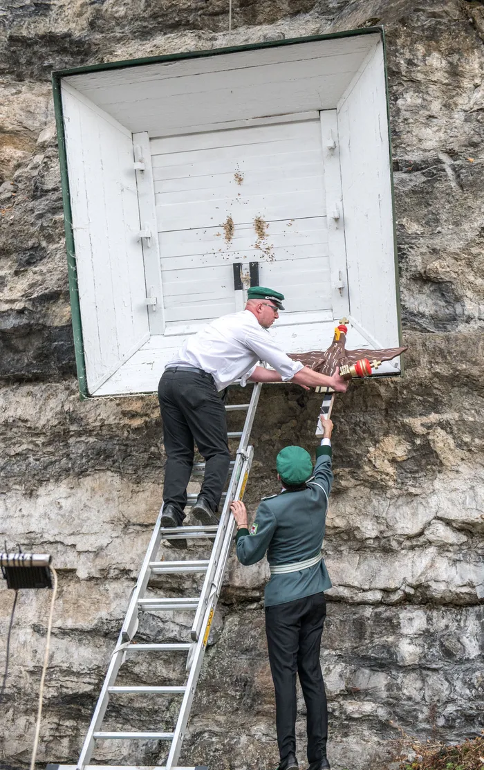 Ein Mann auf einer Leiter hält eine hölzerne Vogelfigur vor eine weiße Holzkiste in einer Felswand, während ein zweiter Mann in Uniform hilft.