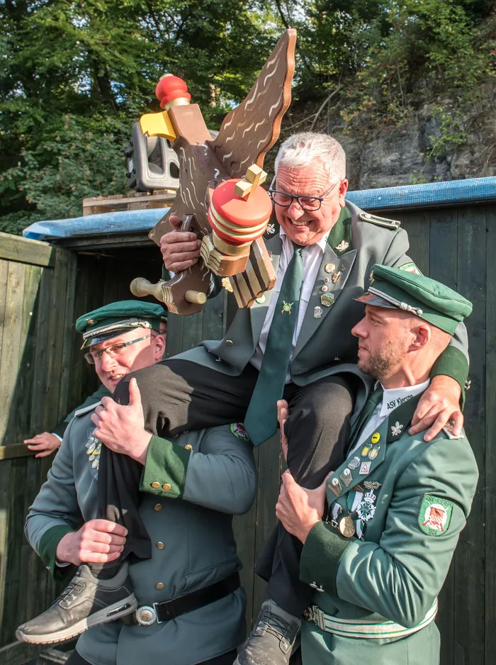 Zwei Männer in grünen Uniformen tragen einen älteren Mann, der eine große Holzfigur eines Adlers hält, beim Schützenfest.