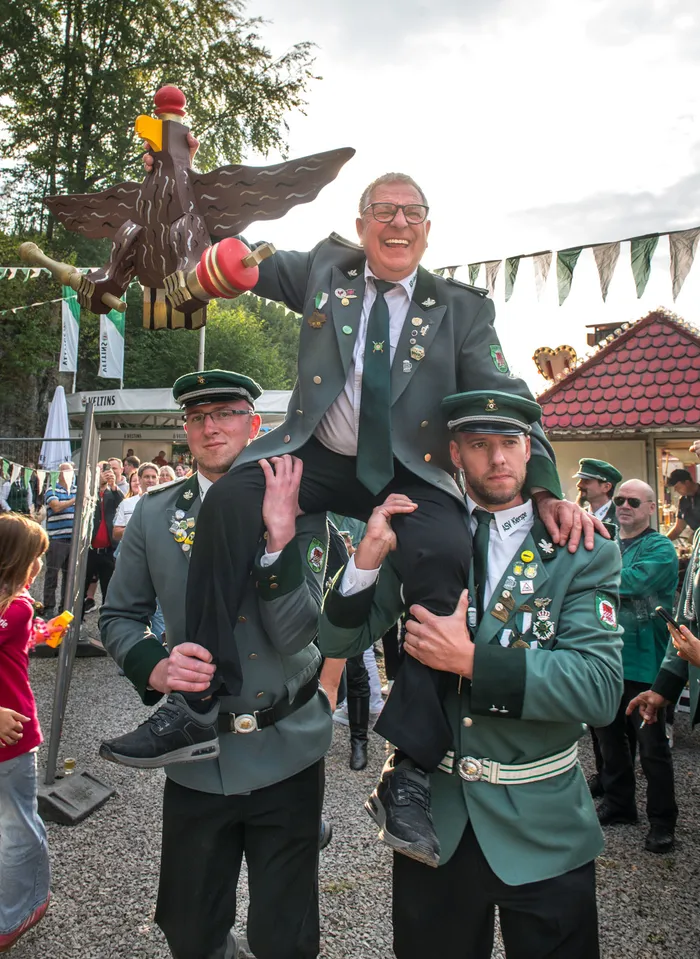 Ein Mann in grüner Uniform wird auf den Schultern zweier Männer getragen und hält eine große Holzadler-Trophäe hoch.