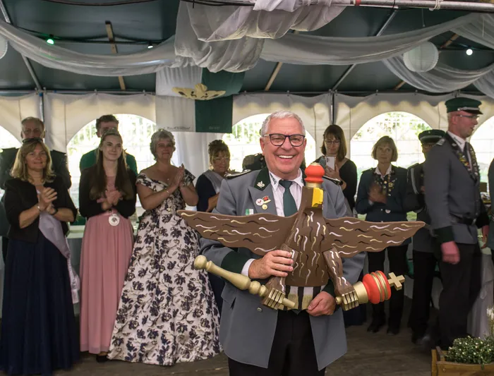 Ein lächelnder Mann in Uniform hält einen großen hölzernen Schützenvogel vor einer Gruppe applaudierender Menschen in einem Festzelt.