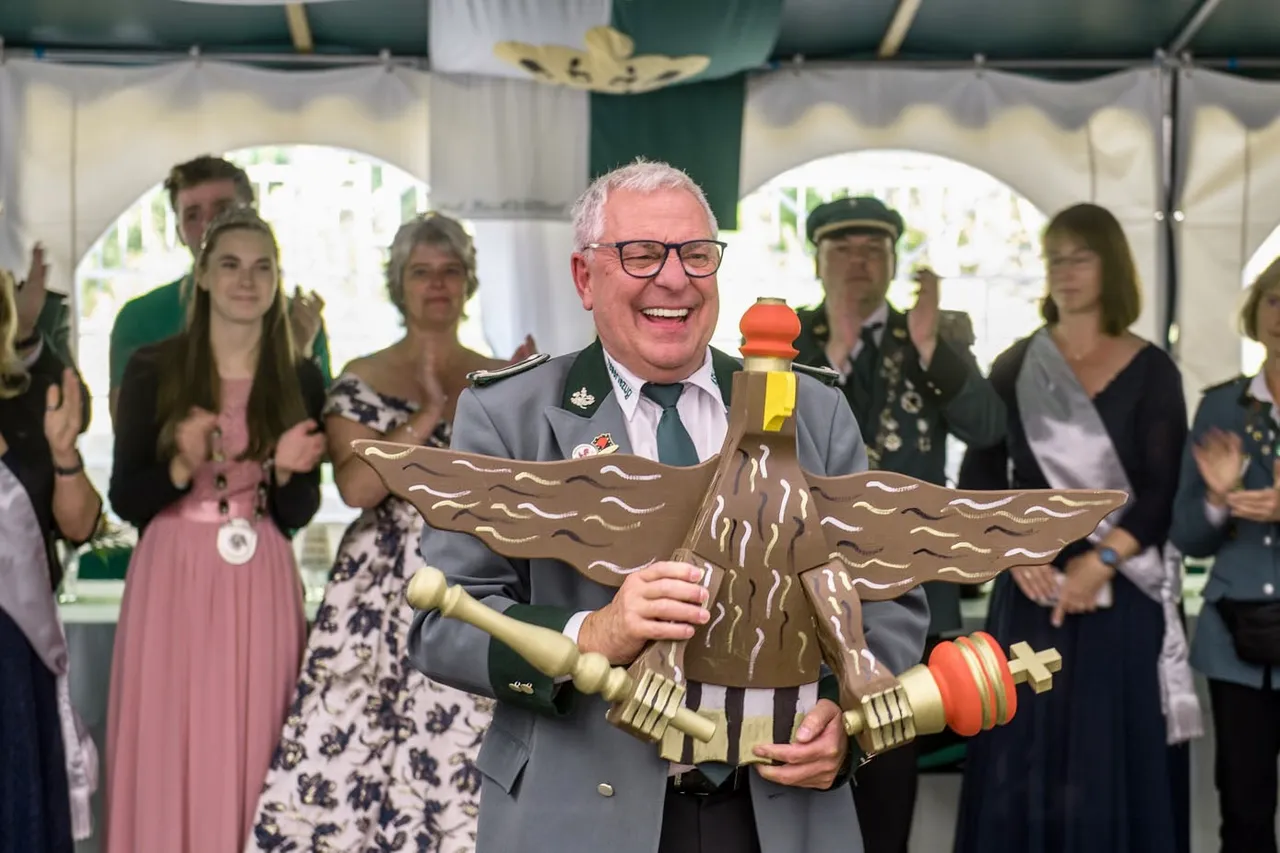 Hugo Mimm lacht und hält eine große, hölzerne Adlerfigur, während Menschen im Hintergrund beim Schützenfest applaudieren.