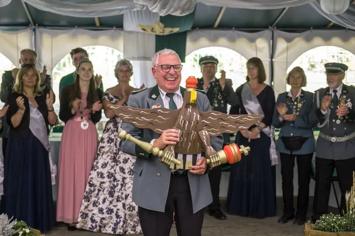 Ein lächelnder Mann in Schützenuniform hält einen großen geschnitzten Holzadler, während im Hintergrund Menschen applaudieren.
