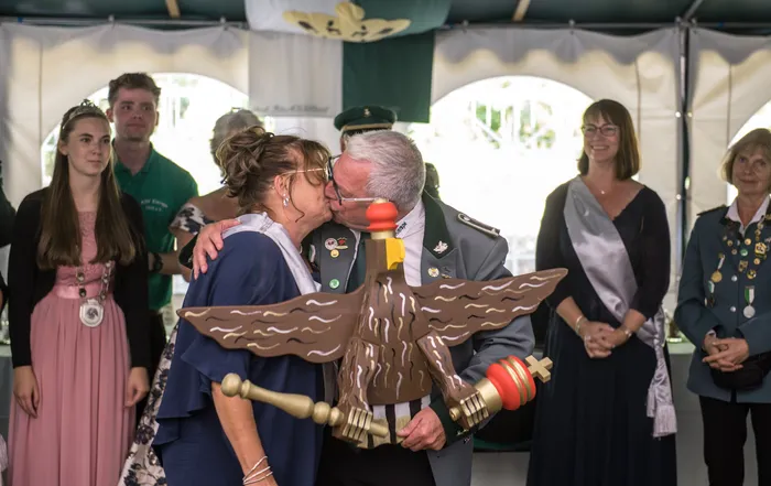 Ein Mann in Uniform küsst eine Frau auf die Wange, im Vordergrund steht eine große Holzfigur eines Adlers.