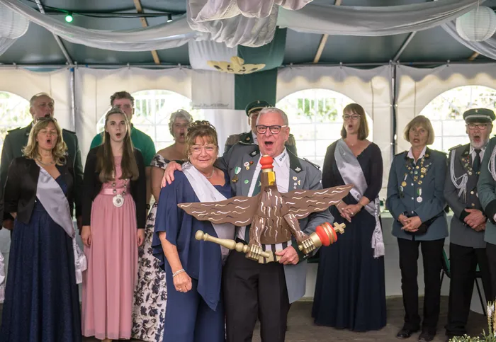 Ein Mann in Uniform präsentiert einen hölzernen Schützenvogel vor einer Gruppe von Menschen in Festkleidung unter einem Festzelt.