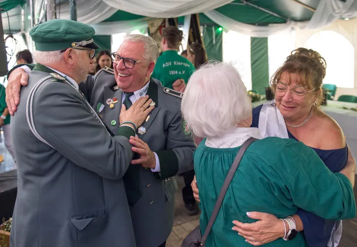 Zwei Männer in grauen Schützenuniformen umarmen sich lachend, während im Vordergrund zwei Frauen sich herzlich begrüßen.