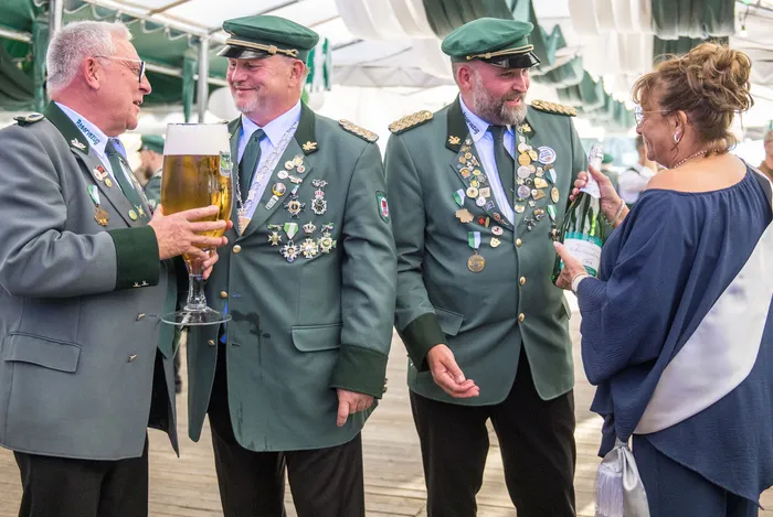 Männer in grünen Schützenuniformen mit Orden stehen bei einem Schützenfest zusammen, einer hält ein Bierglas und eine Frau eine Flasche Sekt.