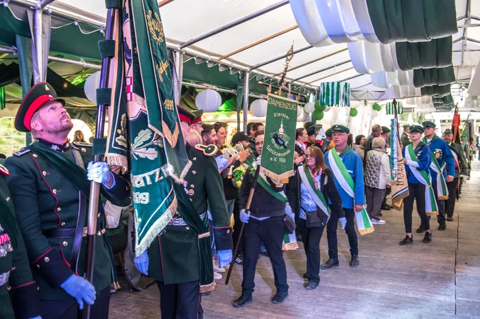 Teilnehmer in Uniformen und mit Fahnen ziehen bei einem Schützenfest in Kierspe in einer Parade unter einem Zeltdach umher.