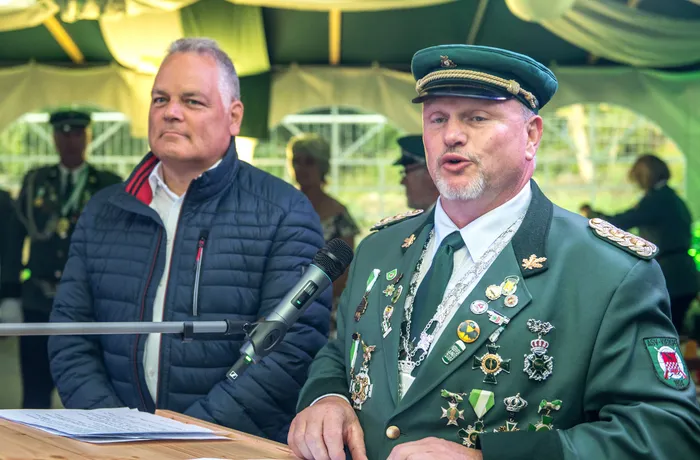 Ein Mann in einer grünen Schützenuniform mit vielen Orden spricht in ein Mikrofon. Neben ihm sitzt ein Mann in einer blauen Jacke.