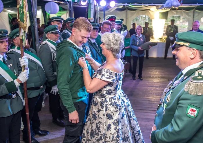 Eine Frau in einem floralen Kleid steckt einem jungen Mann in einem grünen Kapuzenpullover eine Anstecknadel an, umgeben von Männern in grünen Uniformen.