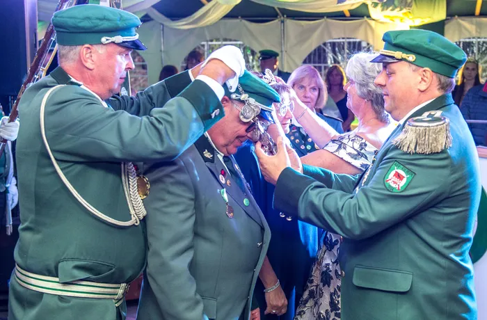 Zwei Männer in grünen Schützenuniformen richten einem dritten Mann in Uniform eine Kopfbedeckung auf den Kopf.