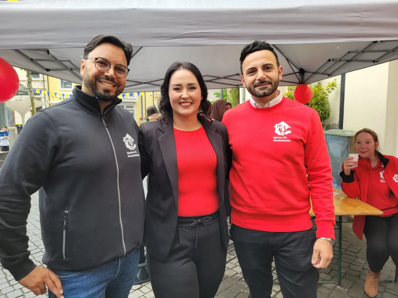 Drei Personen stehen lächelnd vor einem Stand mit roten Ballons. Die Frau in der Mitte und die beiden Männer tragen Kleidung mit dem Logo der „Agentur für Haushaltshilfe“. Im Hintergrund steht ein Pavillon und weitere Personen sind zu sehen.