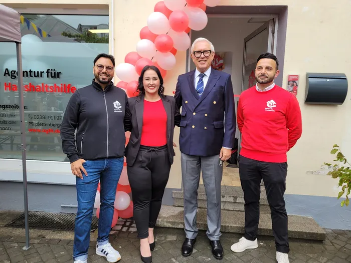 Vier Personen stehen vor dem Eingang einer Agentur für Haushaltshilfe. Die Frau in der Mitte trägt ein rotes Oberteil und eine schwarze Hose, während die anderen drei Personen dunkle Jacken tragen. Im Hintergrund sind rote Luftballons zu sehen.