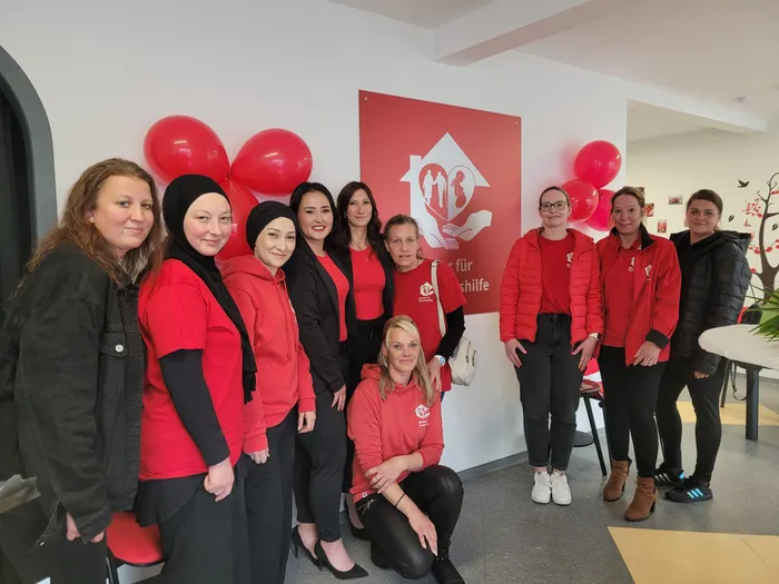 Eine Gruppe von zehn Frauen, die meisten tragen rote Poloshirts mit dem Logo der Agentur für Haushaltshilfe, posiert lächelnd in einem hellen Büro. Im Hintergrund ein rotes Banner mit dem Logo und dem Schriftzug 