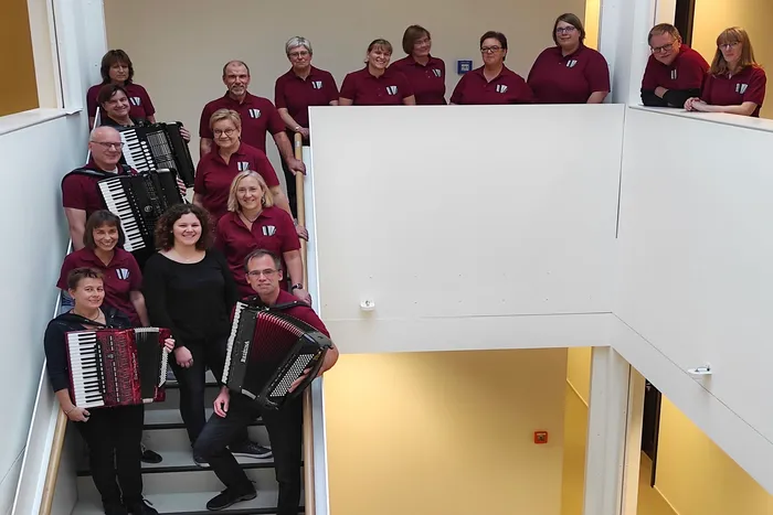 Ein großes Akkordeonorchester in roten Poloshirts posiert auf einer Treppe in einem hellen Gebäude. Einige Mitglieder halten ihre Instrumente vor sich.
