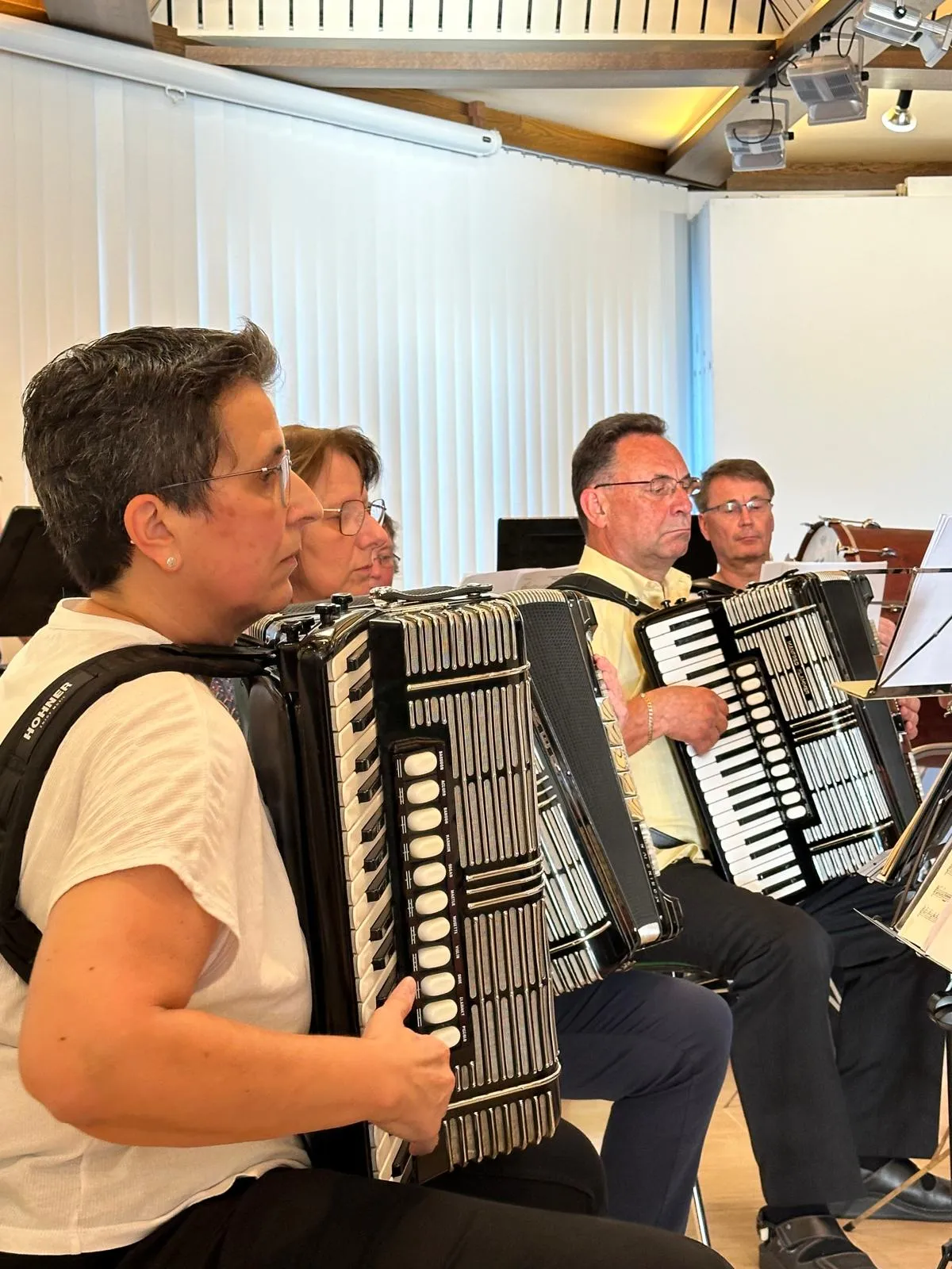 Mehrere Musiker spielen auf Akkordeons in einem hellen Raum, vermutlich bei einem Konzert. Im Hintergrund sind weitere Musiker und Schlagzeug zu sehen.
