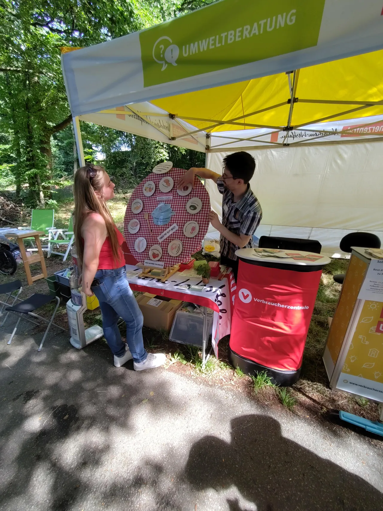 Eine Frau und ein Mann stehen an einem Stand mit dem Logo der Verbraucherzentrale und Informationsmaterialien zum Thema Lebensmittelverschwendung auf einem Stadtparkfest. Im Hintergrund ist ein Zelt mit der Aufschrift 