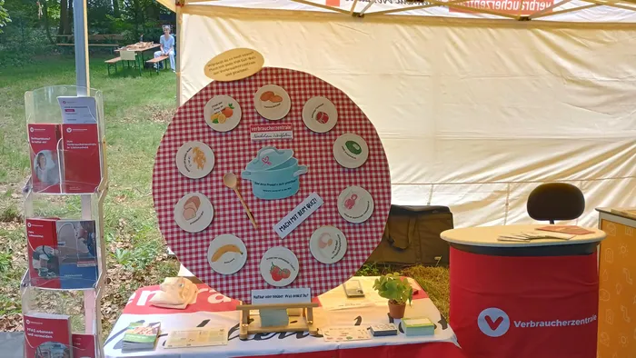 Ein Stand der Verbraucherzentrale NRW auf dem Stadtparkfest in Lüdenscheid informiert über Lebensmittelverschwendung. An einem rot-weiß karierten Hintergrund sind Illustrationen von Lebensmitteln und eine Schüssel mit dem Aufdruck 