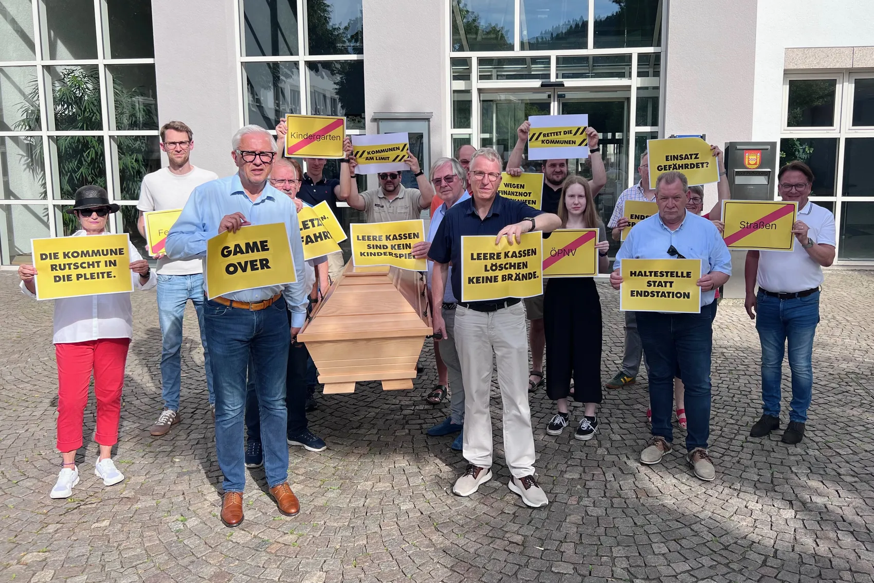 Eine Gruppe von Menschen steht vor einem Rathaus und hält Schilder mit Aufschriften wie 'Die Kommune rutscht in die Pleite' und 'Leere Kassen löschen keine Brände'. Sie demonstrieren gegen die kommunale Finanznot.