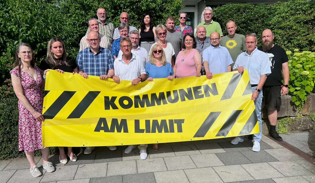 Eine Gruppe von etwa 20 Personen hält ein gelbes Banner mit der Aufschrift „Kommunen am Limit“ vor einem Gebäude. Die Demonstrierenden stehen auf einem gepflasterten Weg vor dem Herscheider Rathaus und protestieren für eine bessere finanzielle Ausstattung der Kommunen.