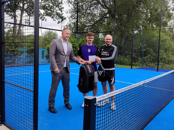 Drei Männer stehen auf einem blauen Padel-Tennisplatz. Links ein Mann im Anzug, in der Mitte und rechts zwei junge Männer, die eine Sporttasche und Padel-Schläger halten.