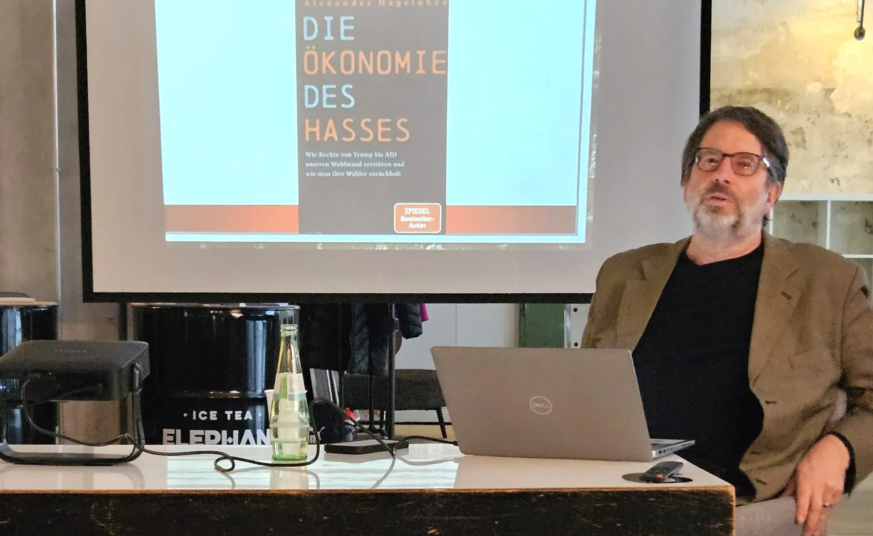 Alexander Hagelüken hält einen Vortrag im Begegnungscafé „Navi“, während auf einer Leinwand der Titel seines Buches „Die Ökonomie des Hasses“ zu sehen ist.