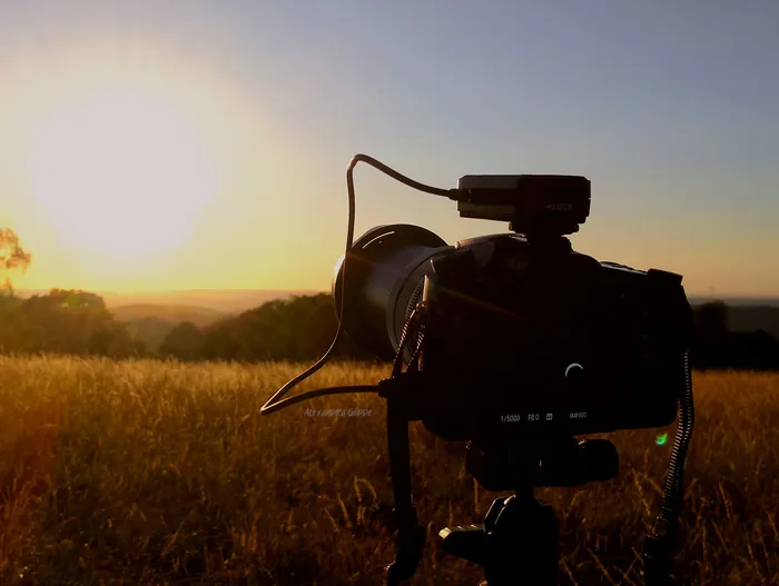 Silhouette einer Kamera auf einem Stativ vor einer tiefstehenden Sonne über einem Feld.