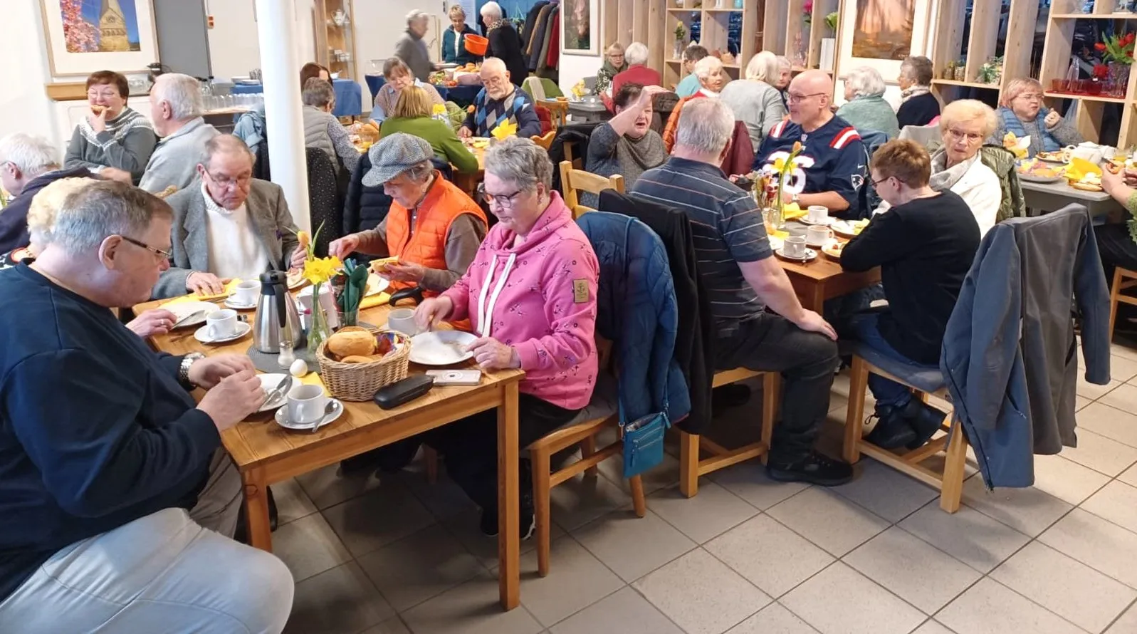 Viele Menschen sitzen an Holztischen in einem hellen Raum und frühstücken. Auf den Tischen stehen Körbe mit Brötchen, Teller mit Aufschnitt und Tassen mit Getränken.