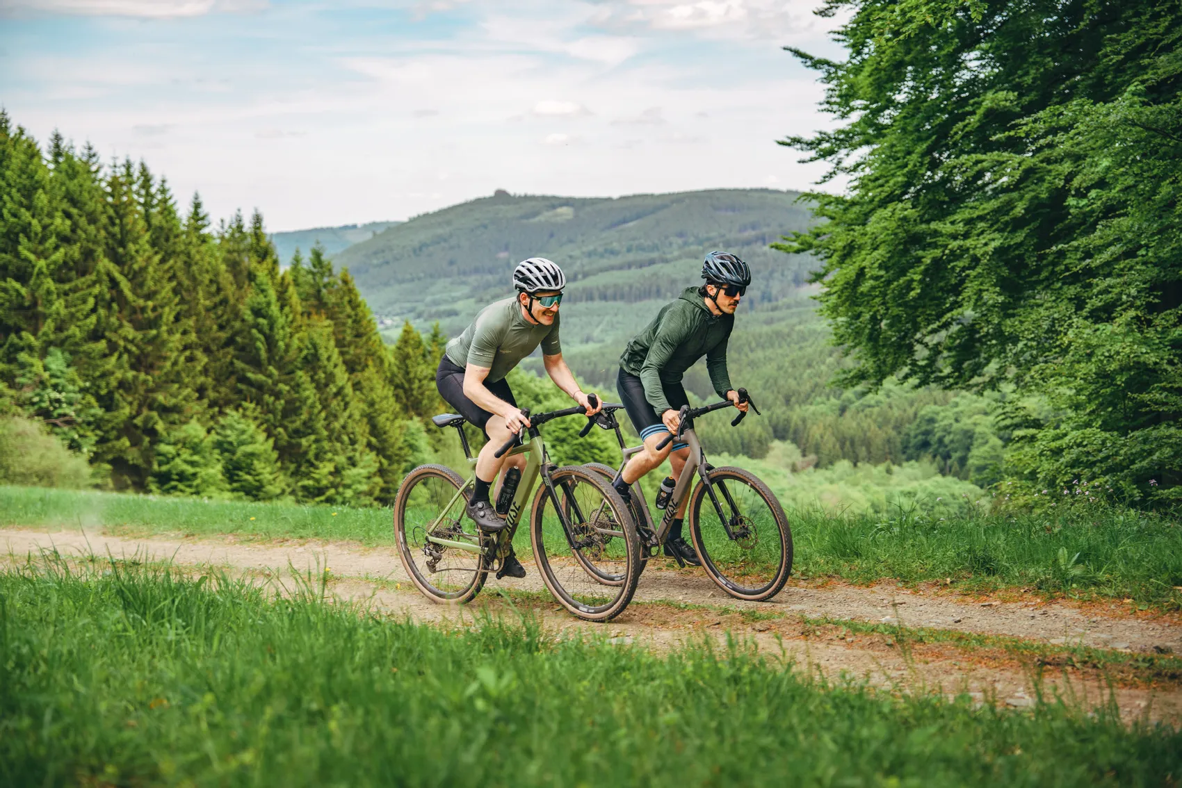 Zwei Männer fahren auf Gravelbikes auf einem Feldweg durch eine grüne Landschaft im Sauerland, im Hintergrund bewaldete Hügel.