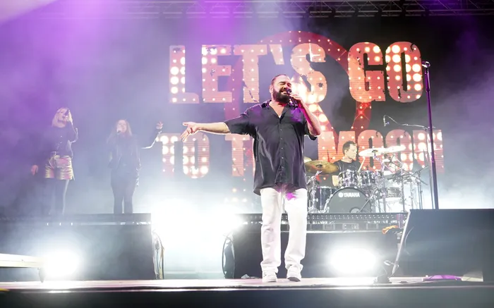 Der Sänger der Band Alphaville singt auf einer Bühne vor einem großen, leuchtenden Schriftzug „LET'S GO TO THE MOON“.