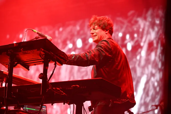 Ein Musiker mit lockigem Haar und Lederjacke spielt an einem Keyboard unter intensiver roter Bühnenbeleuchtung.