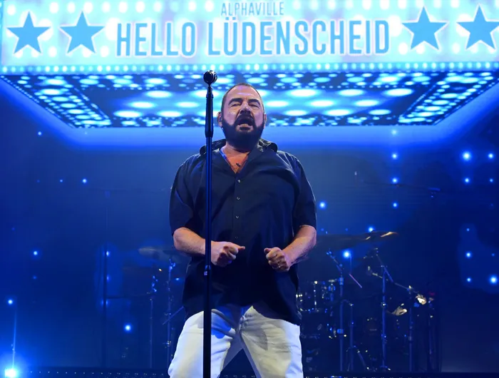 Ein Sänger der Band Alphaville singt auf einer blau beleuchteten Bühne vor einem Schild mit der Aufschrift „ALPHAVILLE HELLO LÜDENSCHEID“.